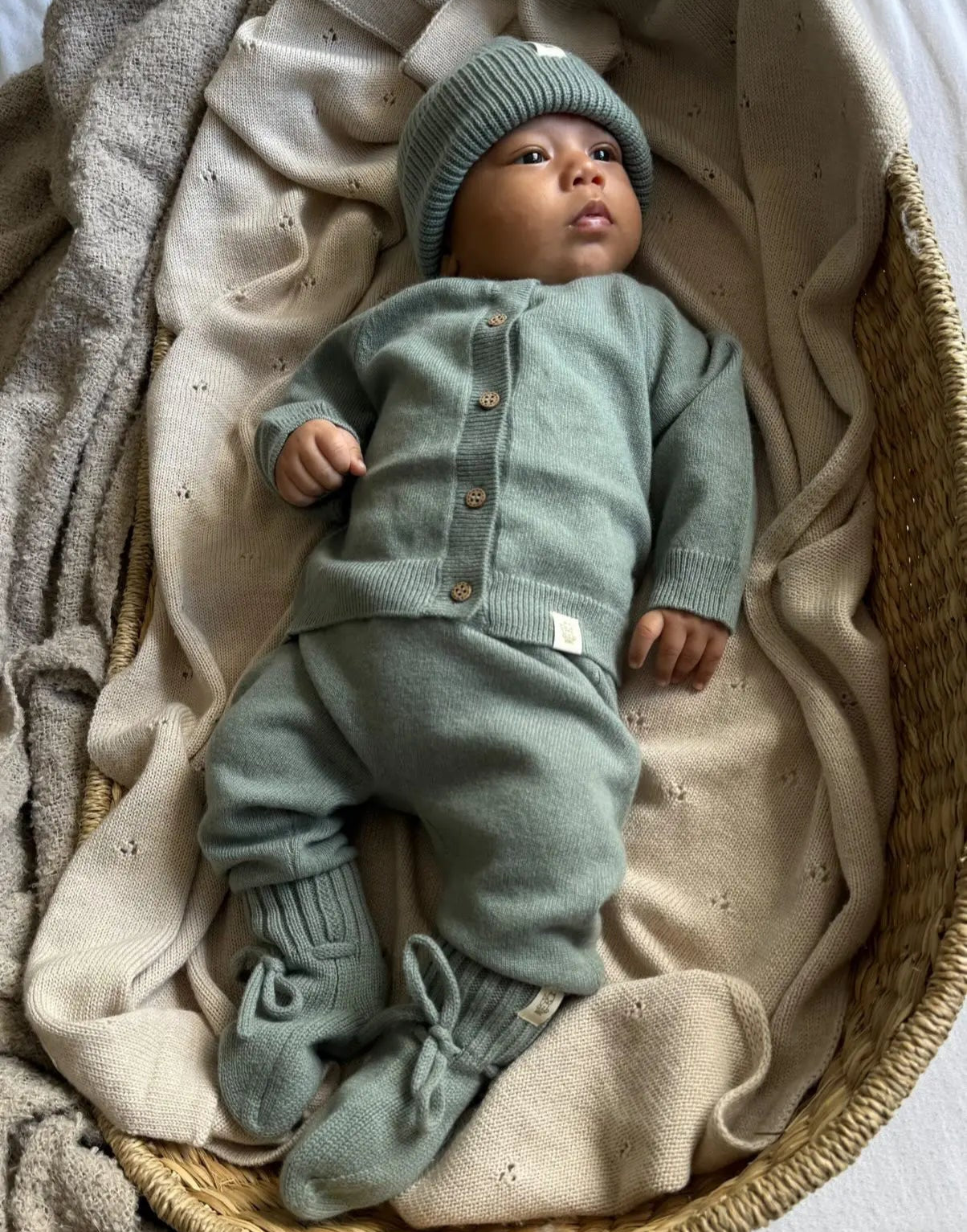 Baby in wollen sage groene Moalie outfit. de baby draagt een mutsje, vestje, broekje en slofjes  uit dezelfde serie merinowol en cashmere kleding collectie.