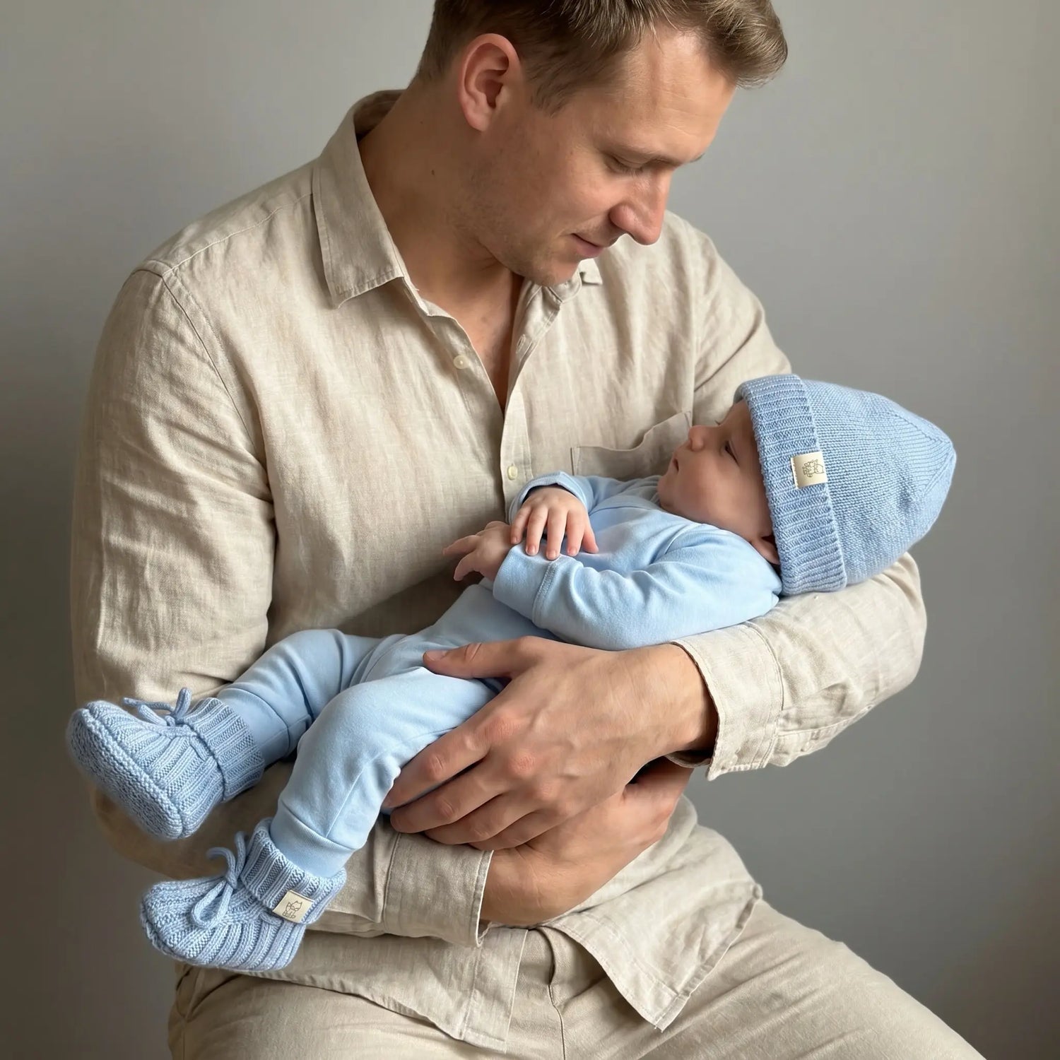 baby gekleed in baby blauwe outfit, betaansden uit een overslag romper en broekje van jersey en een mutsje en slofjes van merinowol en cashmere. De baby ligt in de armen van vader, die hem liefdevol aankijkt. 