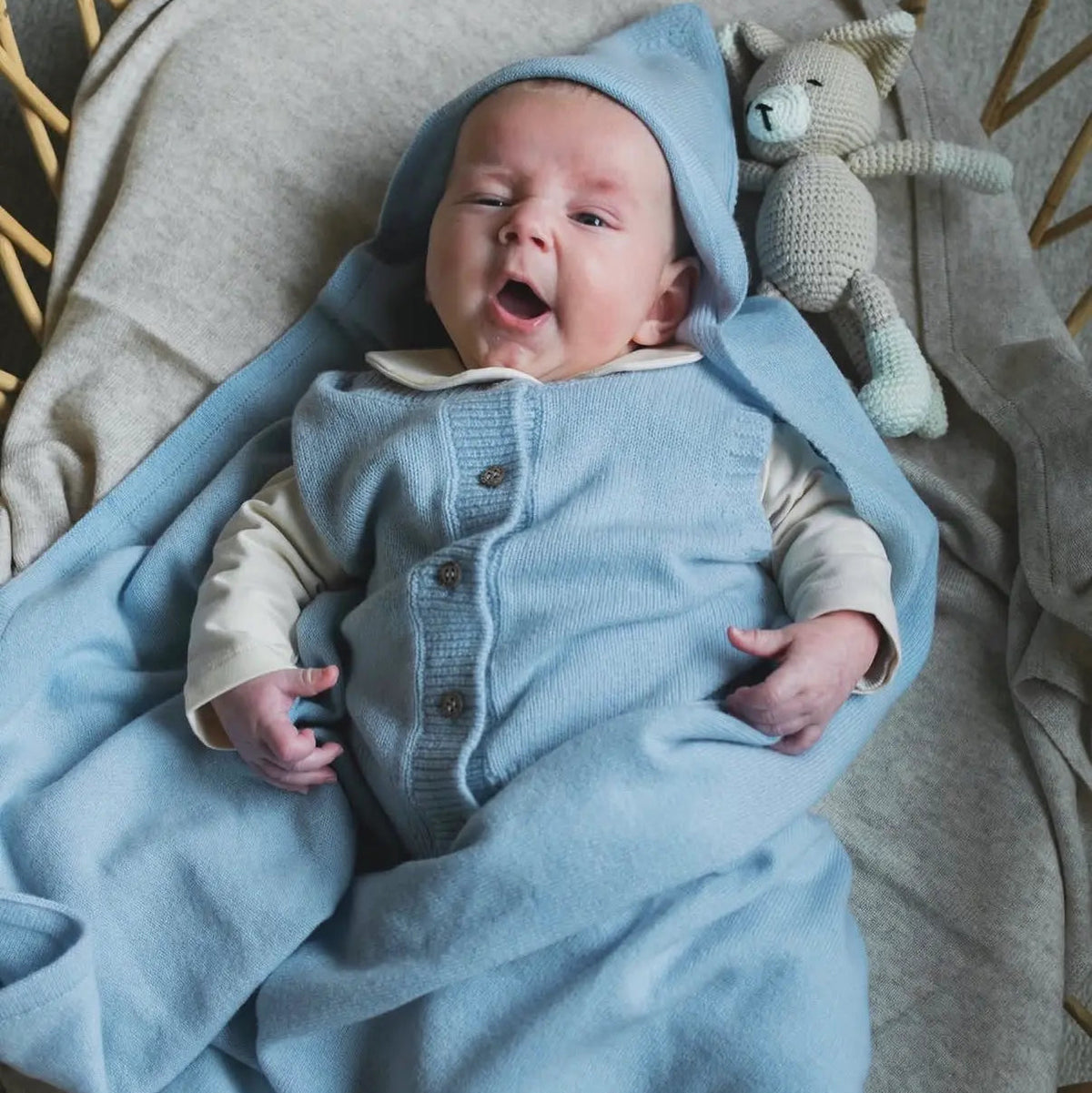 Gapende vrolijke baby in rotan wieg. Gewikkeld in een zacht blauwe deken van merinowol en cashmere. 