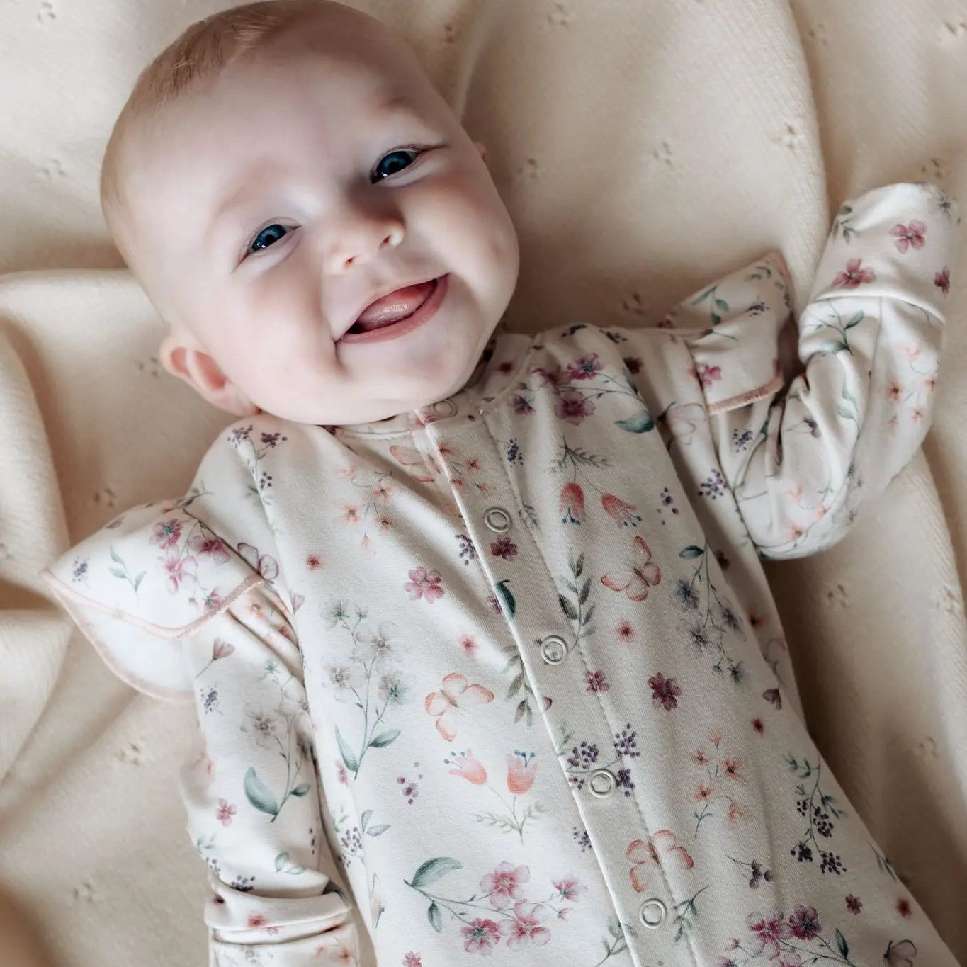 Lachende baby op wollen deken. de baby draagt een boxpakje gemaakt van zacht roze jersey met een print van bloemen en vlinders. Het boxpakje heeft ruffles langs de schouders.
