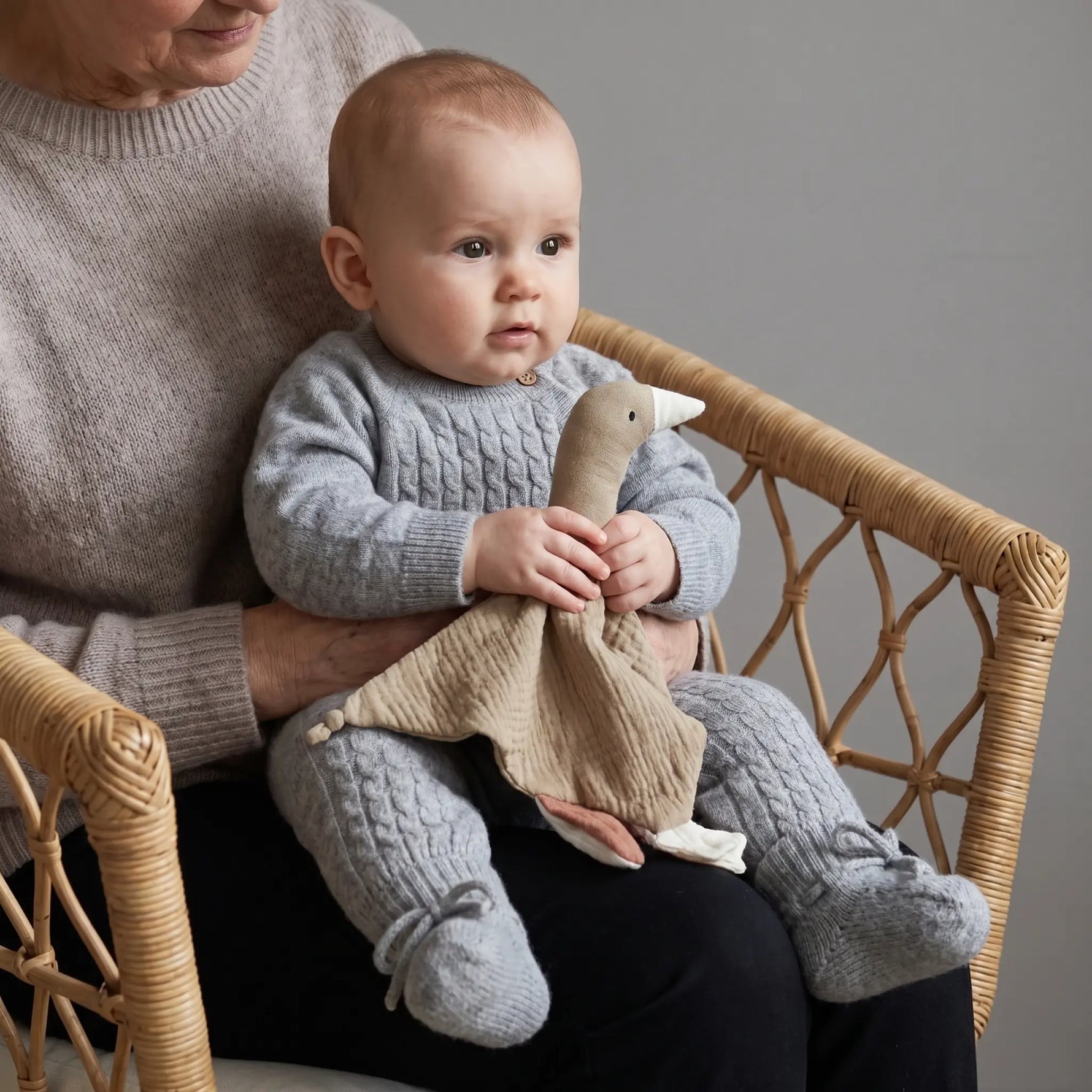 Baby bij oma op schoot in rotan stoel. De baby draagr een grijze boxpakje van merinowol en cashmere en bijpassende slofjes. De baby houdt een knuffeldoekje in taupe kleur vast. Het doekje is in de vorm van een gans en gemaakt van mousseline. 