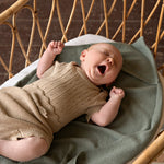 gapende baby in rotan wieg. de baby draagt een gebreid setje van romper en broekje van beige katoen