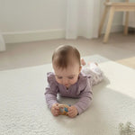 baby spelend in  buikligging op een off white kleed op de grond. De baby draagt een lila kleurige longsleeve met ruches en een gebloemde legging