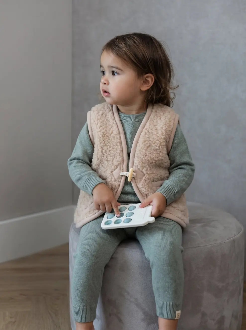 Peuter zittend op grijze krukje gekleed in sage groen pakje van merinowol en cashmere en een beige bodywarmer van merinowol