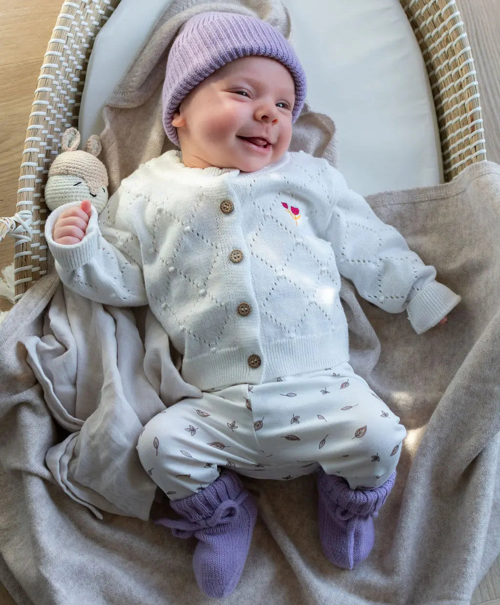 lachende baby in Moalie outfit. Lila kleurig mutsje en slofjes van merinowol en wit gebreid vestje van katoen.