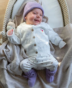 lachende baby in Moalie outfit. Lila kleurig mutsje en slofjes van merinowol en wit gebreid vestje van katoen.