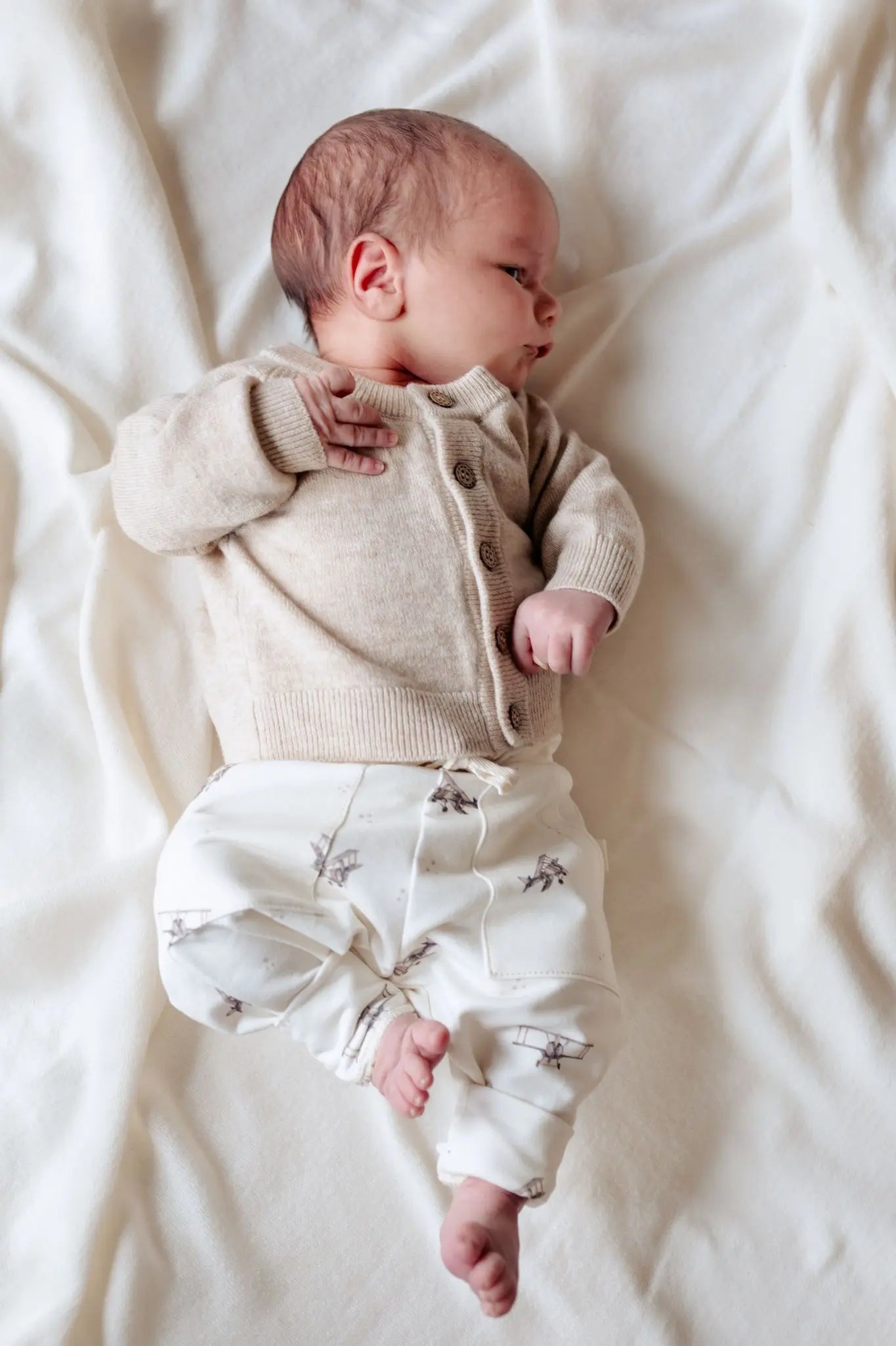Baby liggend op witte deken. De baby draagt een beige vestje van merinowol en een jogger broekje in off white met bruin gekleurde print van vintage vliegtuigjes.