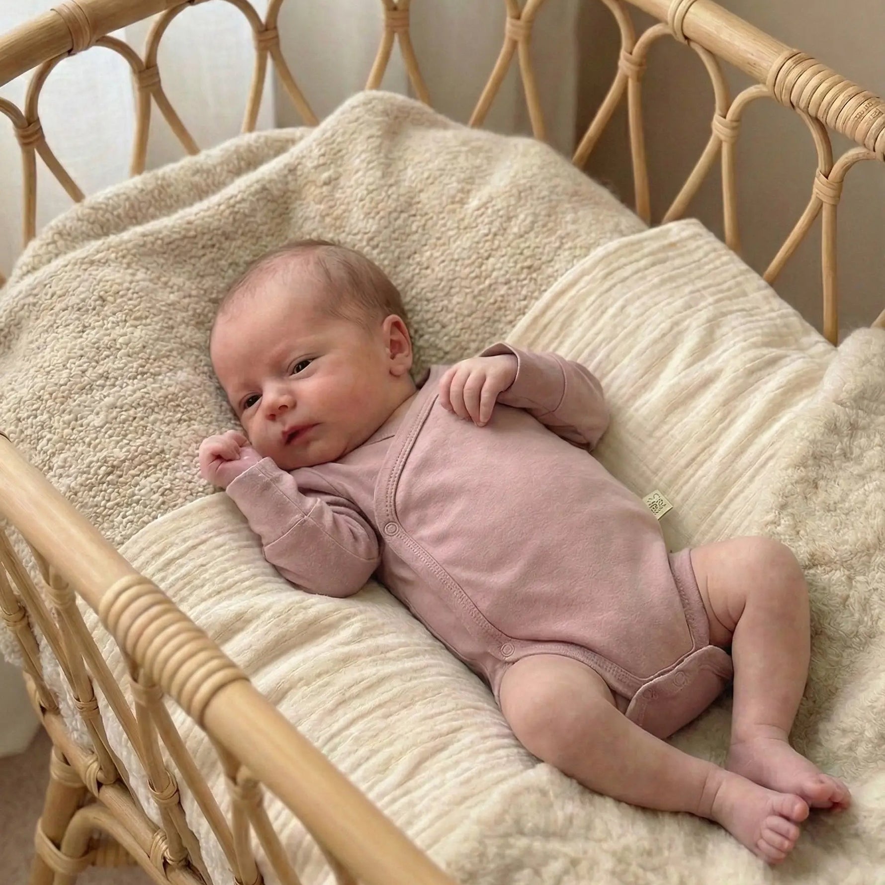 baby liggend in rotan wiegje. De baby draagt een oud roze overslag en meegroeromper van merinowol en bamboe. 
