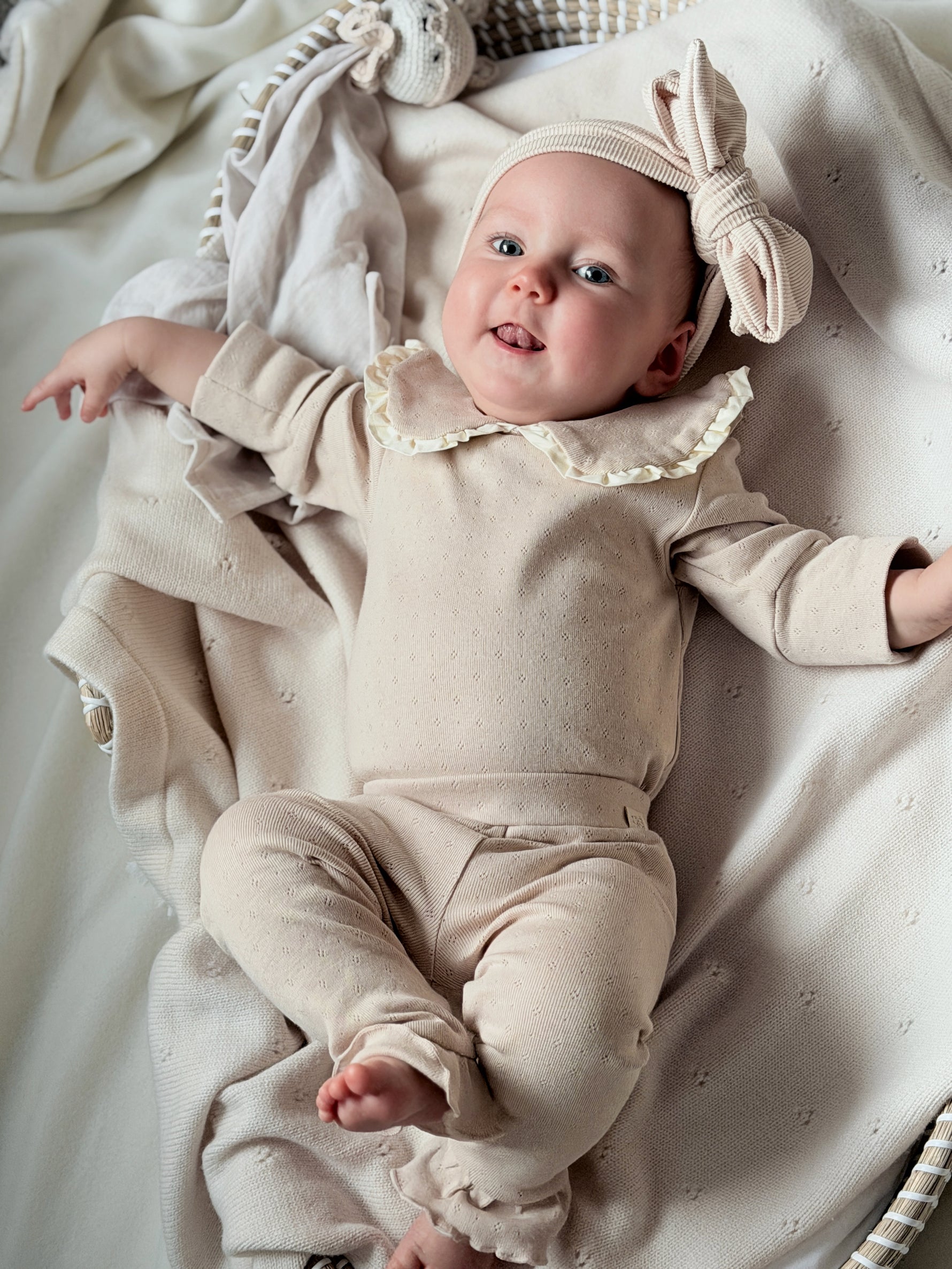Baby liggend op crèmekleurig kleed. De baby draagt een Pointelle setje van lichtbeige jersey. De set bestaat uit een romper met grote geschulpte kraag en een legging met ruffle onderaan de pijpjes. De baby draagt een haarband met grote strik in de kleur beige 