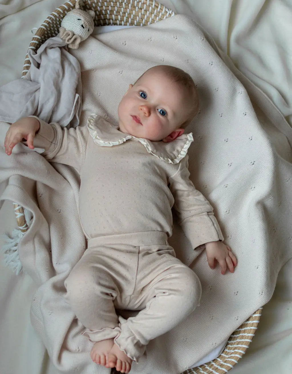 Baby op wollen deken liggend. de baby kijkt in de lens en draagt een pakje gemaakt van zacht beige pointelle jersey. de romper heeft een klassiek franse kraag 