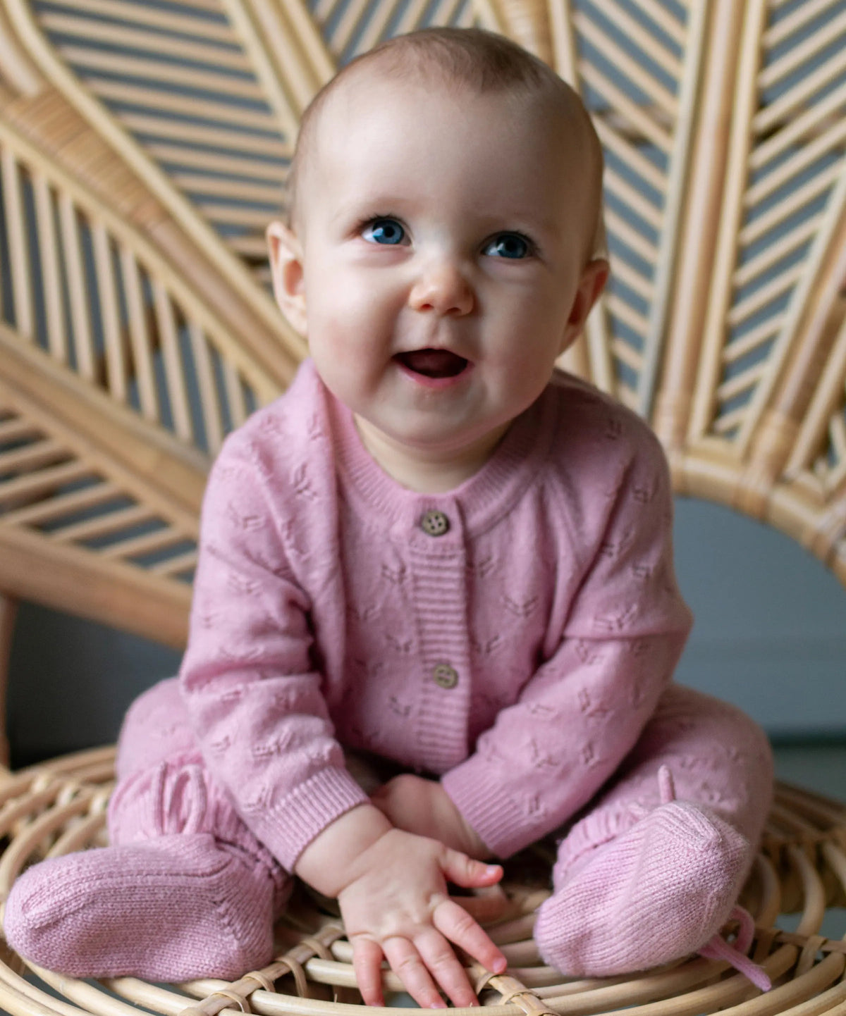 Baby zittend in rotan stoel. De baby draagt  een roze boxpakje van merinowol en cashmere en bijpassende roze wollen slofjes