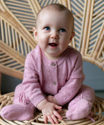 Baby zittend in rotan stoel. De baby draagt  een roze boxpakje van merinowol en cashmere en bijpassende roze wollen slofjes