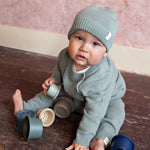 baby zittend op de grond in een sage groene merinowollen outfit van Moalie bestaande uit een mutsje, sweater en broekje