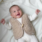 lachende baby in creme kleurige wollen boxpakje en merinowollen beige gilet 
