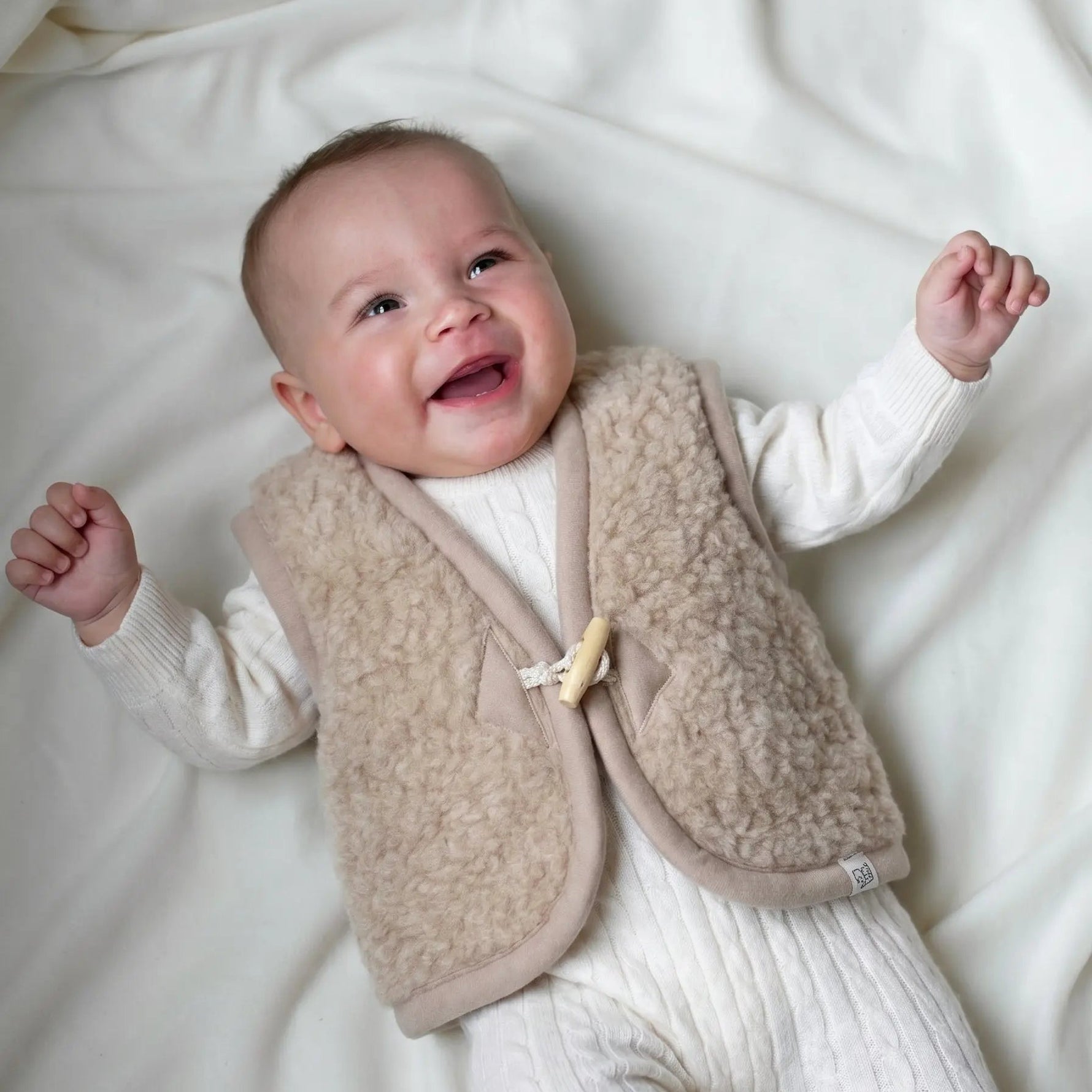 lachende baby in creme kleurige wollen boxpakje en merinowollen beige gilet 