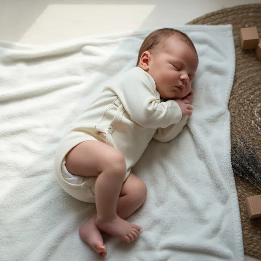 baby in ecru kleurige wollen romper van merinowol en bamboe, liggen op een witte deken op de grond.
