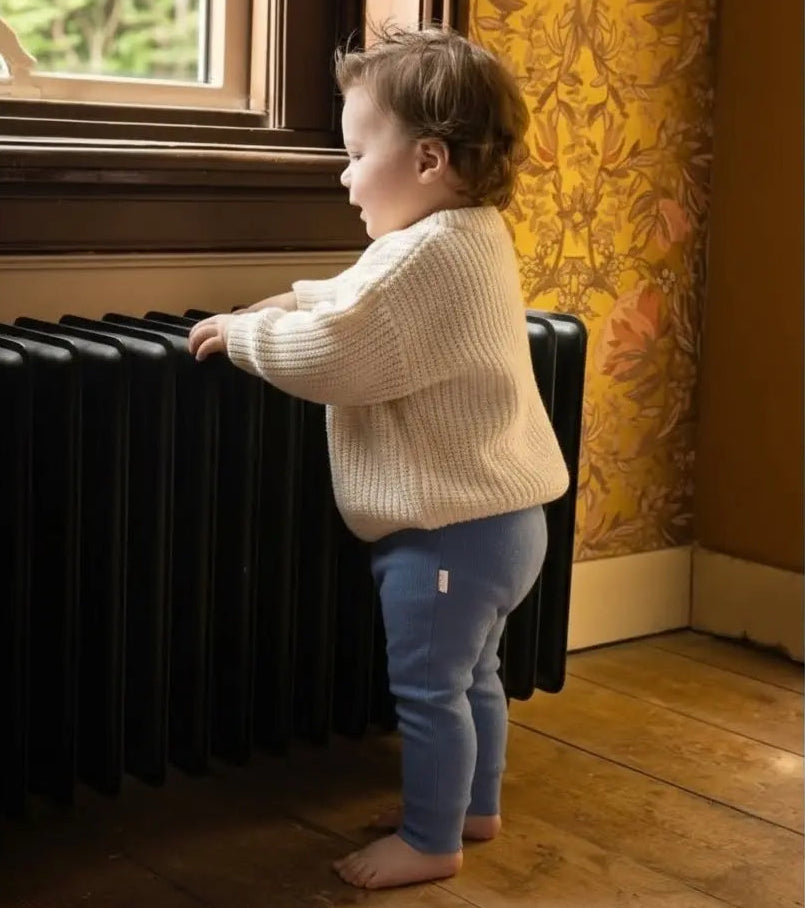 baby in vintage look woonkamer staande aan zwarte radiator. De baby draagt een blauw broekje van gebreide katoenen stof en een katoenen big knit sweater n creme kleur.