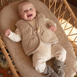 baby in rootan wieg liggned op katoenen beige deken gekleed in merinowollen beige gilet, wollen creme kleurig boxpakje en babyslofjes Rice Flower 