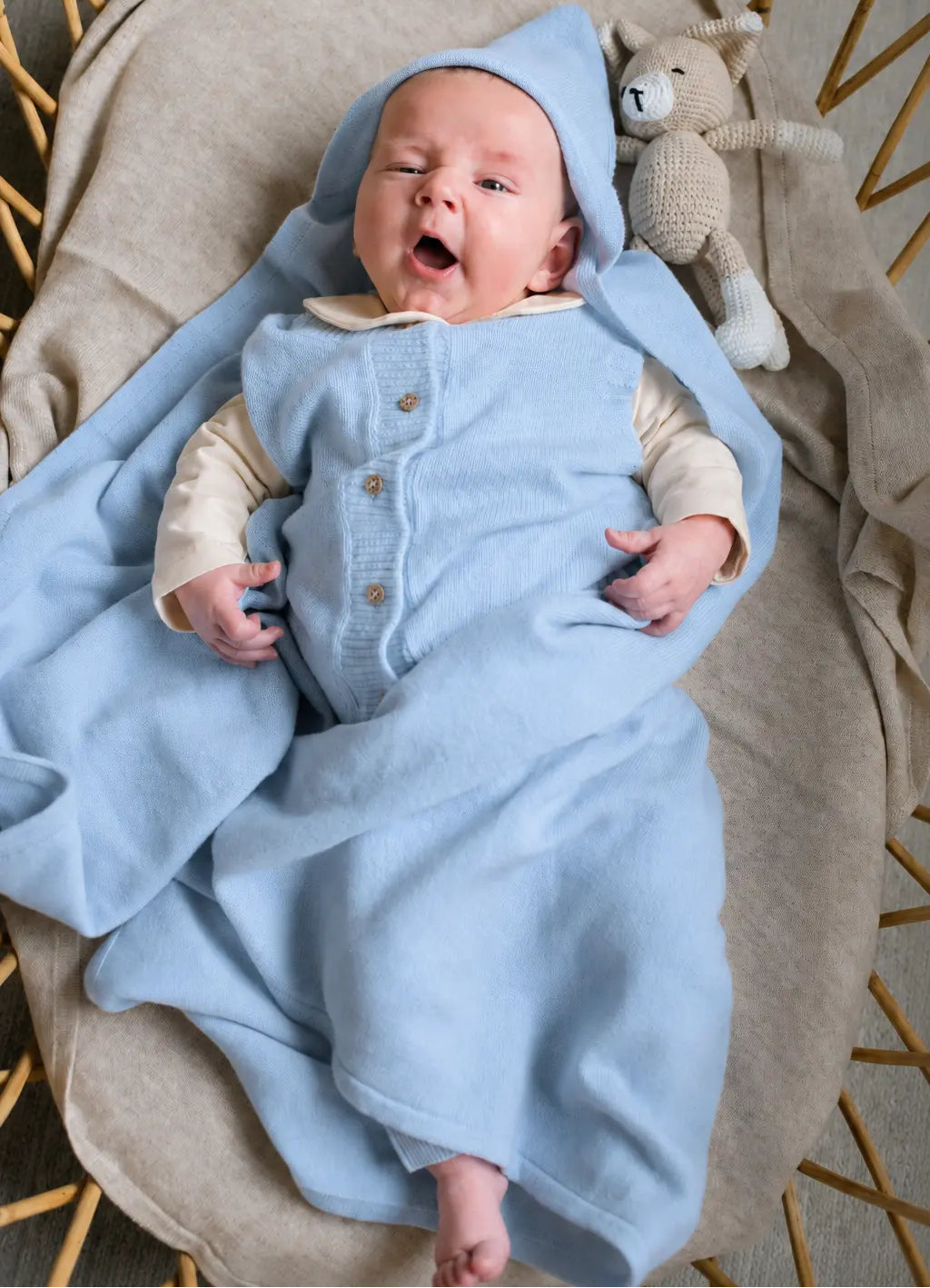 Gapende vrolijke baby in rotan wieg. Gewikkeld in een zacht blauwe deken van merinowol en cashmere. 