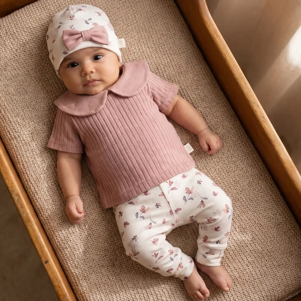 Baby in houten wiegje gekleed in het Petal geboortepakje van Moalie – oud roze ribstof shirtje, Petal Legging met bloemenprint en bijpassende mutsje met strik