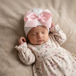 sleeping newborn on beige blanket. The baby is wearing a white newborn beanie wit pink bow and cotton light pink dress with flower pattern