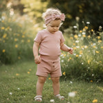 Peuter staand in gras en gekleed in een t-shirt, korte broek en haarband van oud roze biologisch katoen