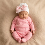 newborn in pink babygrow with footies and a white newborn beanie with pink satin bow