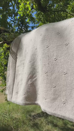 video van merinowollen pointelle deken in licht beide zandkleur. De deken hngt over een tak in een boomgaard en wordt door de wind zacht en en weer gewiegd