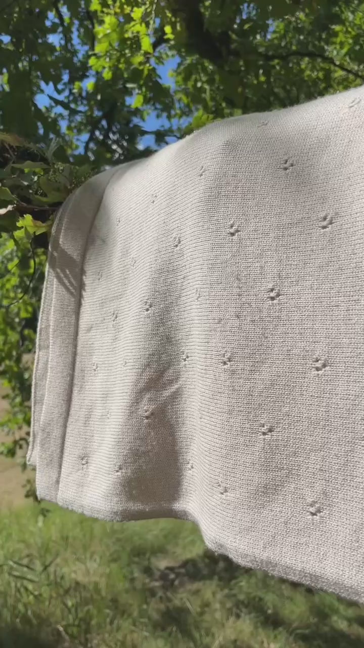 video van merinowollen pointelle deken in licht beide zandkleur. De deken hngt over een tak in een boomgaard en wordt door de wind zacht en en weer gewiegd