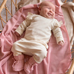 lachende baby op roze merinwollen deken liggend in off white katoenen pakje met ruffles en roze merinowollen baby slofjes 