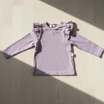 Lila kleurige longsleeve met ruffles rond de schouders op een houten vloer, verlicht door de zon
