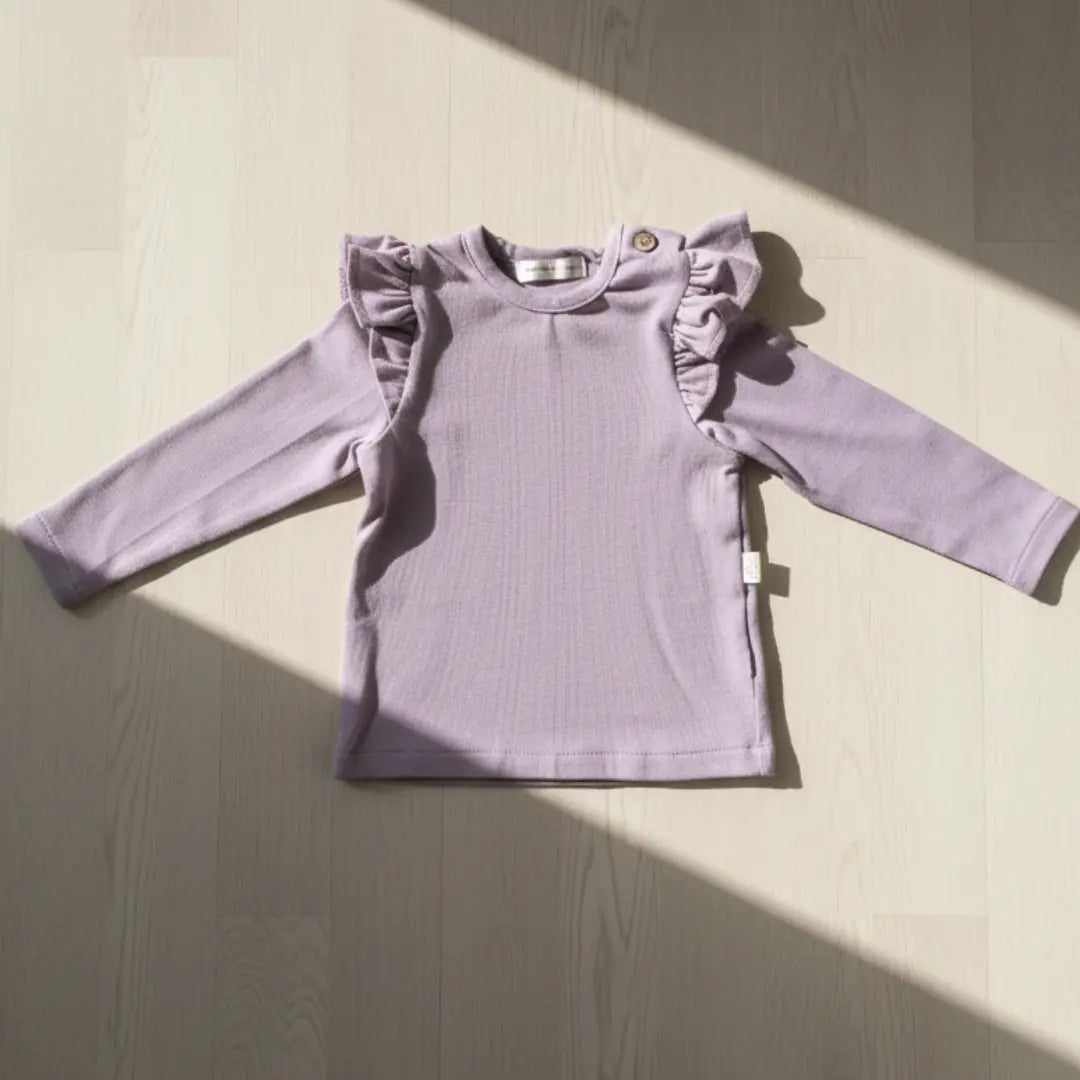 Lila kleurige longsleeve met ruffles rond de schouders op een houten vloer, verlicht door de zon