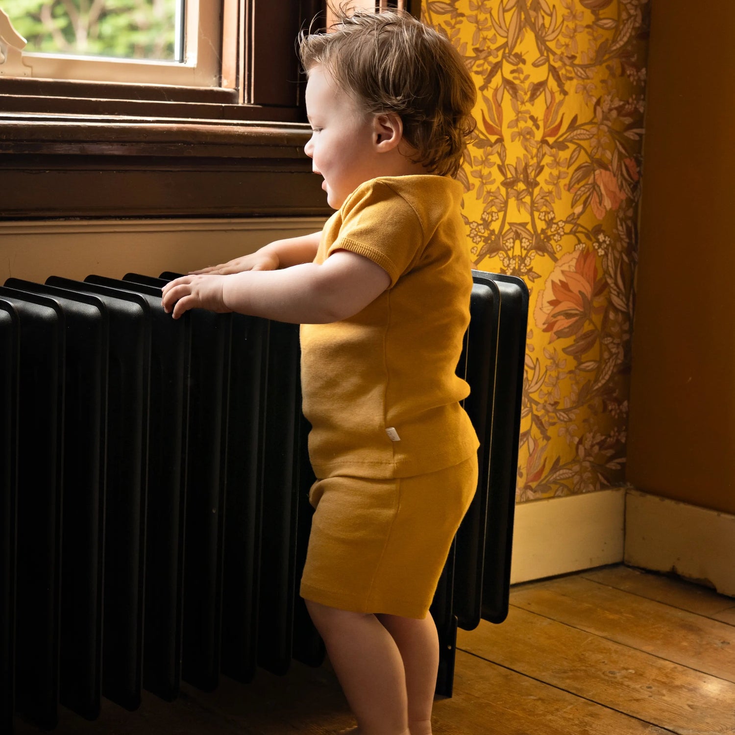 baby staand aan vinatge radiator in een tweedelig baby en peuter setje van biologisch katoen in okergeel 
