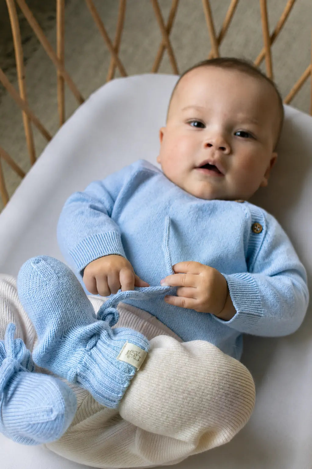 baby in rieten wiegje in een lichtblauwe romper , creme kleurig broekje en lichtblauwe slofjes van merinowol en cashmere van Moalie 