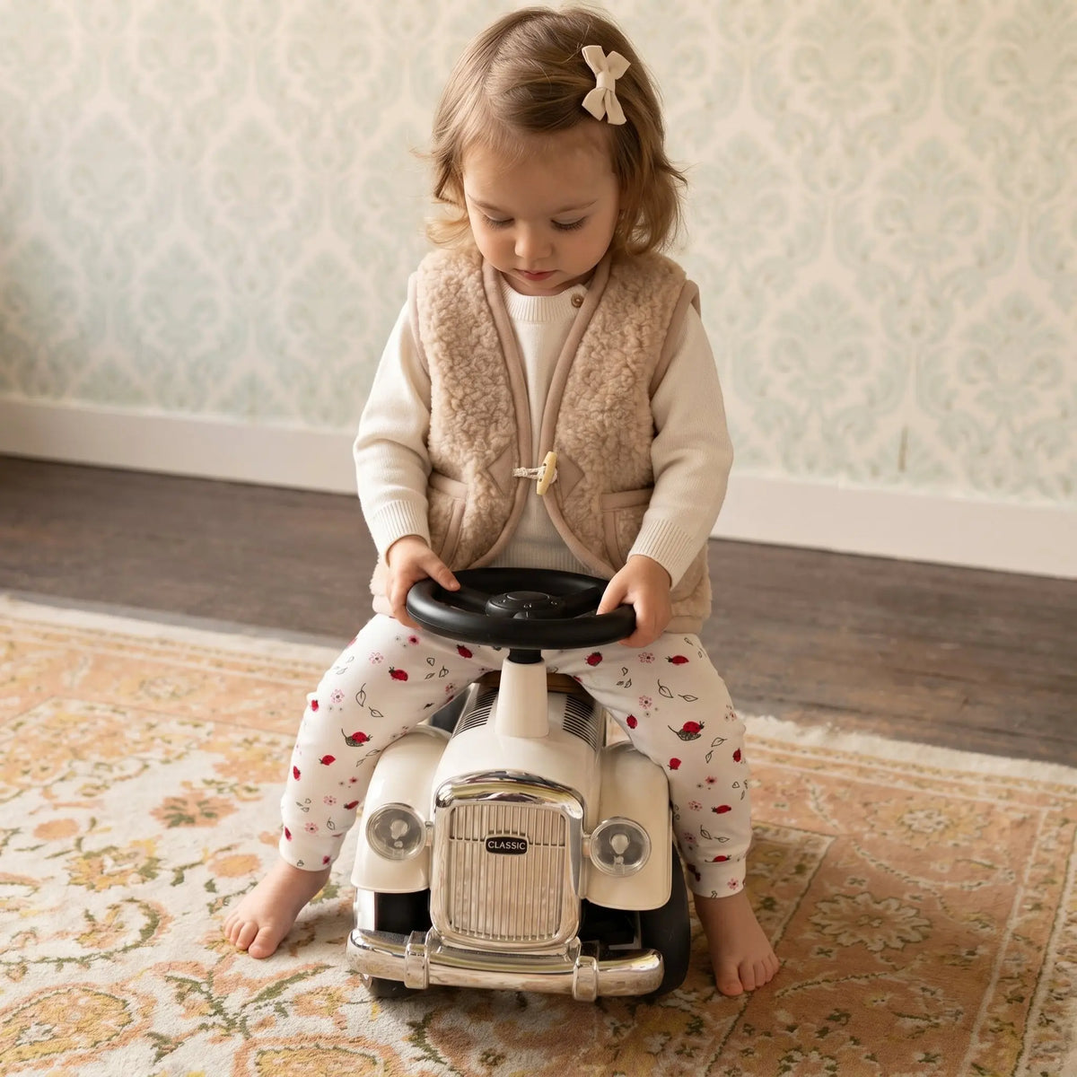 peuter zittend op speelgoed auto. Het meisje draagt een legging met print van lieveheersbeestjes, een creme kleurige merinowollen trui en een beige gekleurde merinowollen bodywarmer.